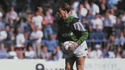 A football goalkeeper in a green kit prepares to kick the ball during a match, with a crowd in the background.