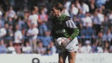 A football goalkeeper in a green kit prepares to kick the ball during a match, with a crowd in the background.