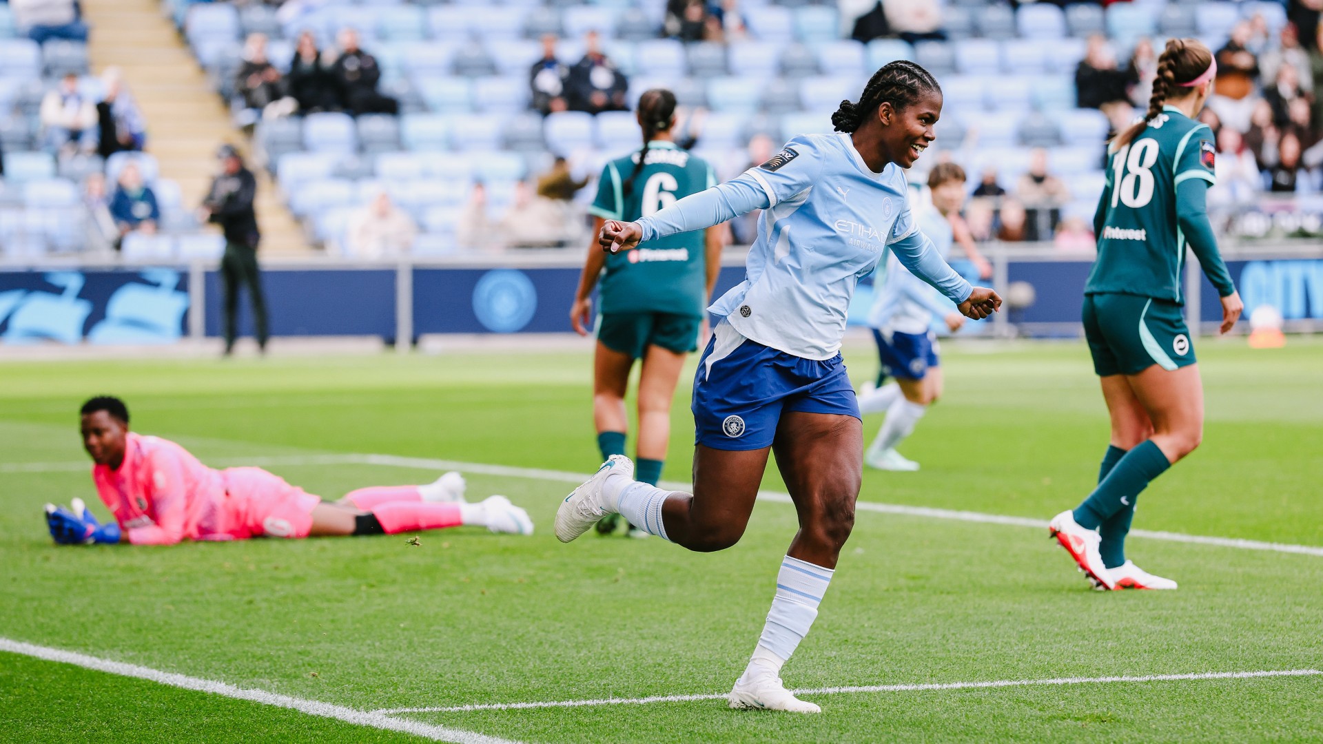 A football player in a light blue and white kit celebrating a goal on the field. Other players and a goalkeeper in a pink kit are visible in the background.