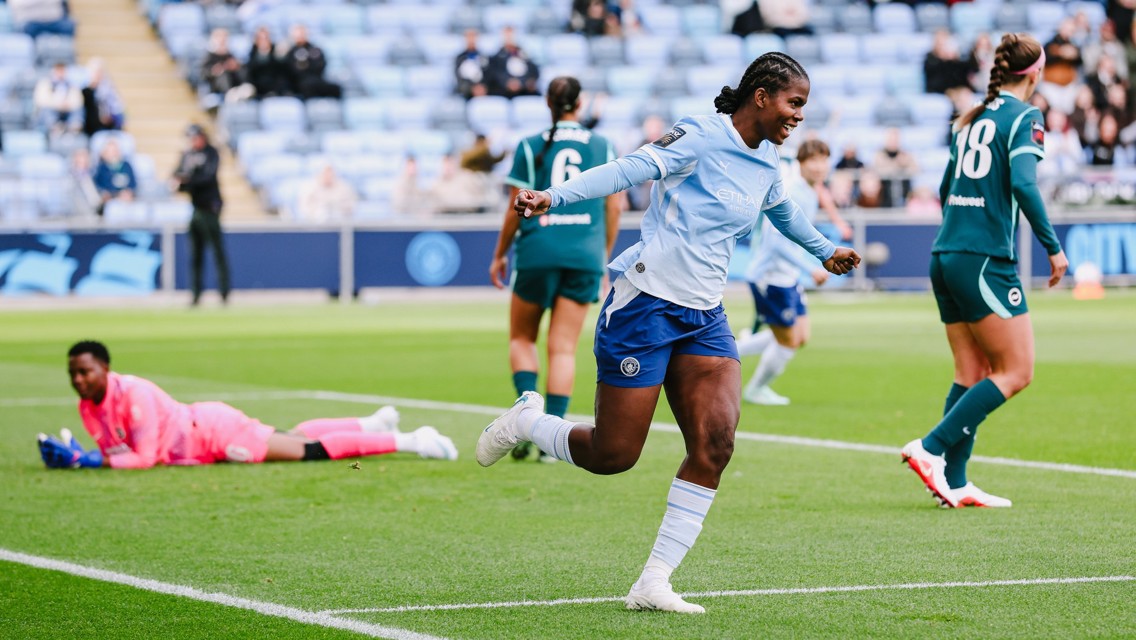 A football player in a light blue and white kit celebrating a goal on the field. Other players and a goalkeeper in a pink kit are visible in the background.