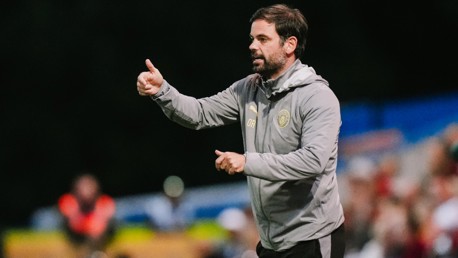 A man wearing a grey jacket with a logo gestures a thumbs up while standing on a sports field.