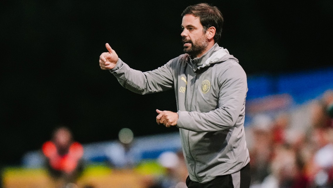 A man wearing a grey jacket with a logo gestures a thumbs up while standing on a sports field.