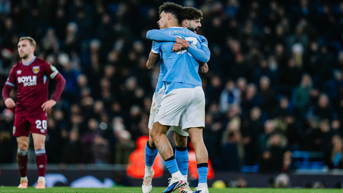 Two football players in light blue jerseys are hugging, celebrating a moment on the field. A player in a claret jersey is visible in the background. The scene is set in a crowded stadium.