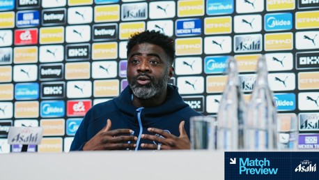 A person speaking at a press conference with a blurred face, Manchester City branding and multiple sponsor logos in the background.