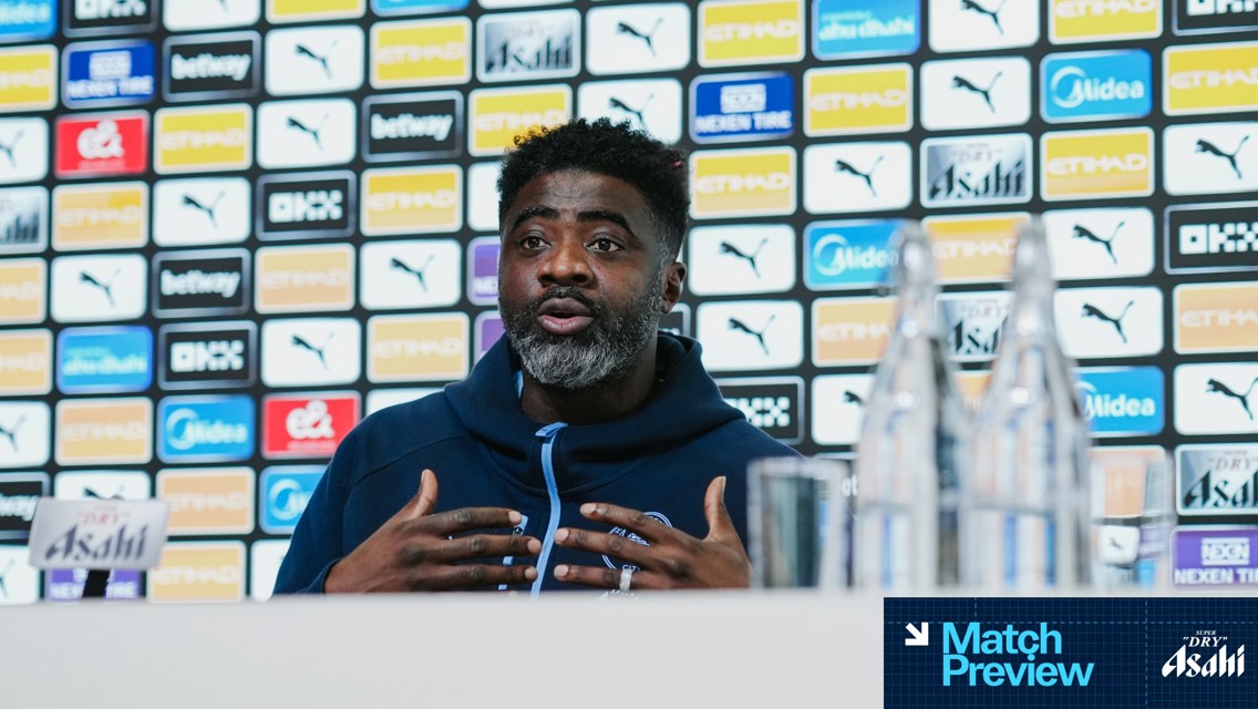 A person speaking at a press conference with a blurred face, Manchester City branding and multiple sponsor logos in the background.