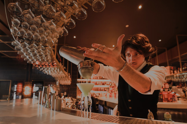 A bartender is skillfully pouring a green cocktail into a glass using a shaker and strainer at a bar with a stylish interior.