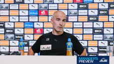 A man in a press conference, seated in front of a backdrop filled with Manchester City sponsors like Puma and Etihad. He has two bottles of Lucozade on the table with a microphone.