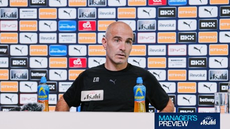 A man in a press conference, seated in front of a backdrop filled with Manchester City sponsors like Puma and Etihad. He has two bottles of Lucozade on the table with a microphone.