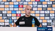 A man in a press conference, seated in front of a backdrop filled with Manchester City sponsors like Puma and Etihad. He has two bottles of Lucozade on the table with a microphone.