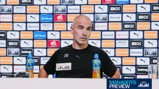 A man in a press conference, seated in front of a backdrop filled with Manchester City sponsors like Puma and Etihad. He has two bottles of Lucozade on the table with a microphone.