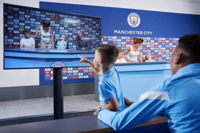 A young boy wearing a Manchester City jersey points at a screen showing him and his family in the Manchester City press room.