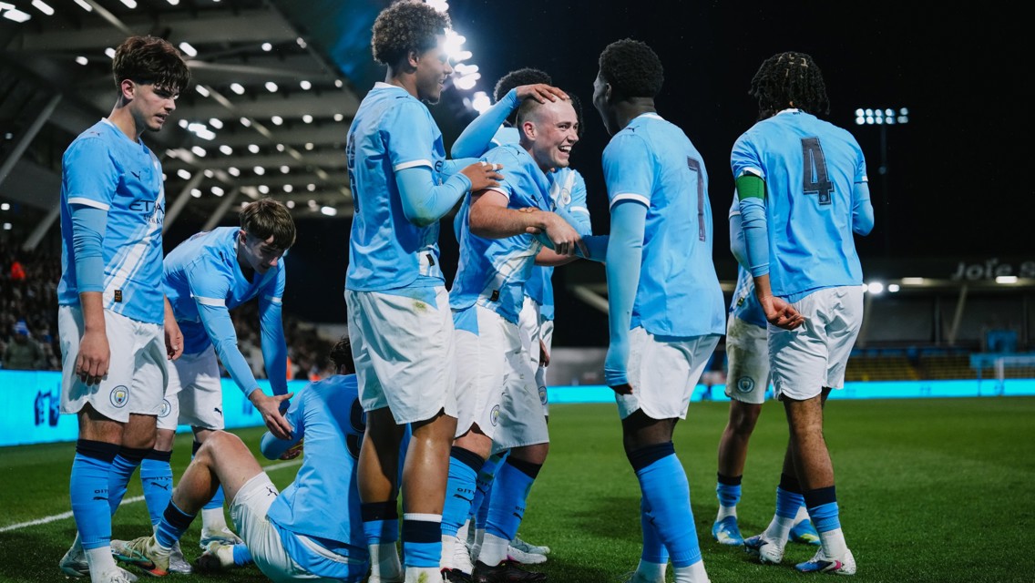 Manchester City football players wearing blue jerseys are gathered on the field celebrating during a night match.