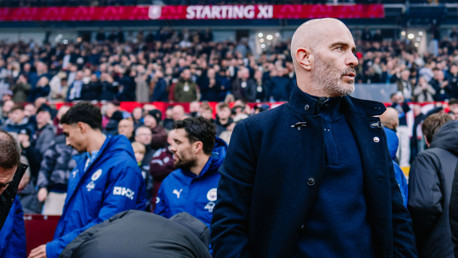 A coach in a dark coat stands near the sideline before a game. Behind him are players wearing blue jackets and a crowd in the background. 'Starting XI' is displayed on a sign above.