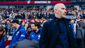 A coach in a dark coat stands near the sideline before a game. Behind him are players wearing blue jackets and a crowd in the background. 'Starting XI' is displayed on a sign above.