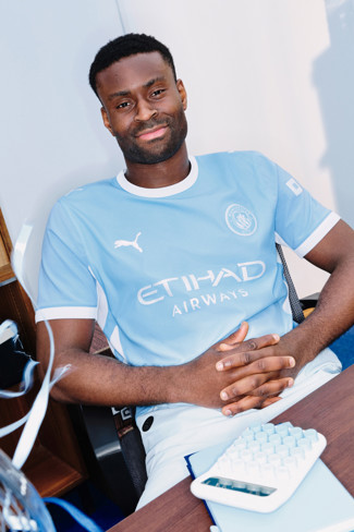 A person wearing a Manchester City jersey sitting at a desk with a keyboard and notebook visible.