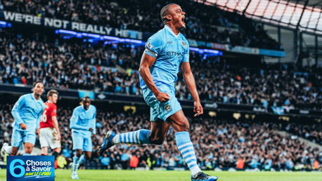Soccer player wearing a Manchester City jersey celebrates during a match at a packed stadium. A mix of Manchester City and opposing team players can be seen in action.