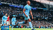 Soccer player wearing a Manchester City jersey celebrates during a match at a packed stadium. A mix of Manchester City and opposing team players can be seen in action.