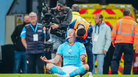 A football player in a Manchester City kit is sitting cross-legged on the field in a celebratory pose, with cameras and crew members nearby.