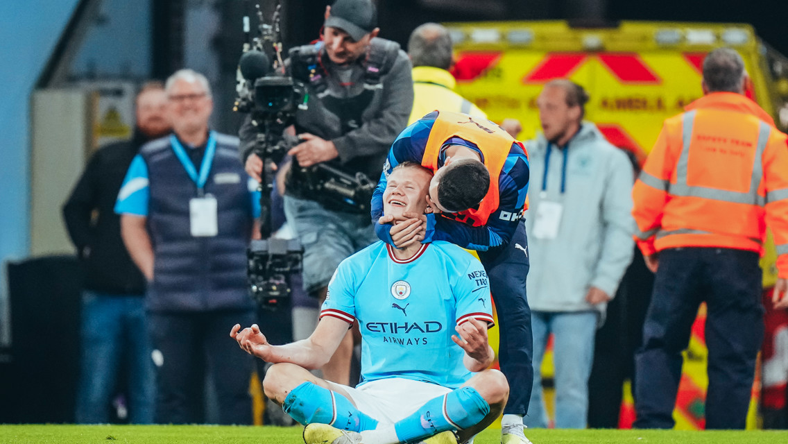 A football player in a Manchester City kit is sitting cross-legged on the field in a celebratory pose, with cameras and crew members nearby.
