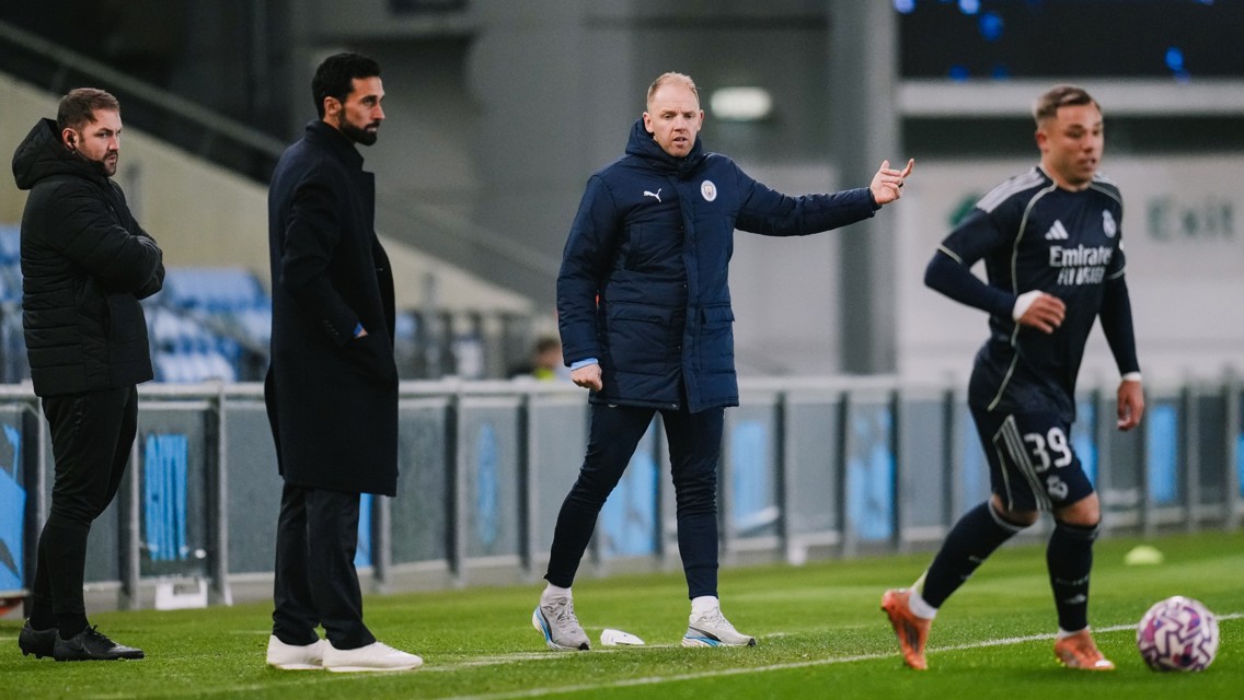 Four individuals stand on the sideline during a soccer match. One person appears to be gesturing, while another seems focused on the field. A player wearing a dark kit with the number 39 is also visible.