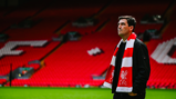 A man wearing a black jacket is standing in a stadium, with a red and white scarf featuring the Liverpool FC logo.