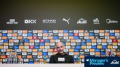 Press conference setup with assorted sponsor logos and branding behind a blurred individual. Brands include Puma, Etihad, OKX, Asahi, etisalat, and Midea. Bottles and beverages on the table labeled 'Asahi' and 'Powerade.'