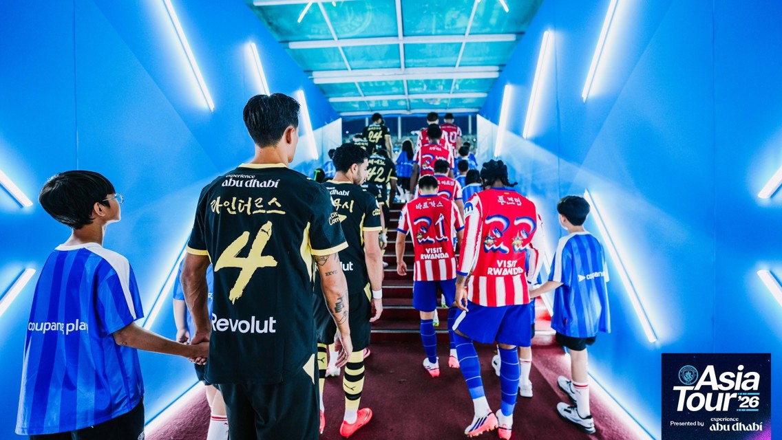 Football players wearing jerseys with team names in Korean are walking up a lit tunnel to enter the field. The image features the logo 'Asia Tour '26' sponsored by Abu Dhabi.