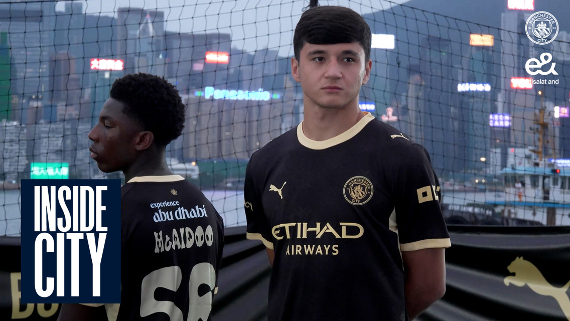 Two men in Manchester City jerseys stand outdoors with a cityscape in the background, marked with 'Inside City.'