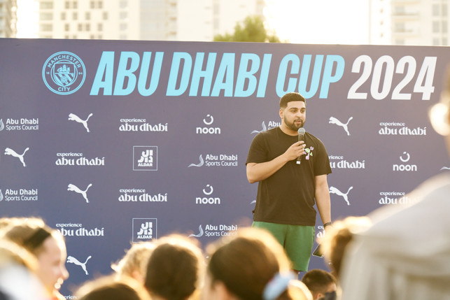 An event speaker is addressing the crowd during the Abu Dhabi Cup 2024, organized by Manchester City. Multiple sponsors and logos are visible on the backdrop including Puma, Abu Dhabi Sports Council, experience Abu Dhabi, noon, and Aldar.