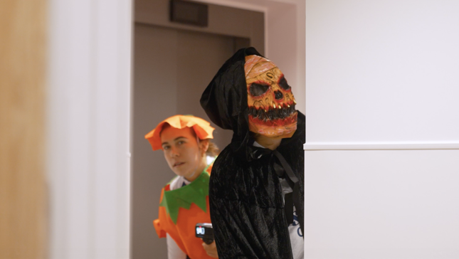 Two people in Halloween costumes peeking around a corner; one in a pumpkin outfit, the other wearing a skull mask with a black hood.