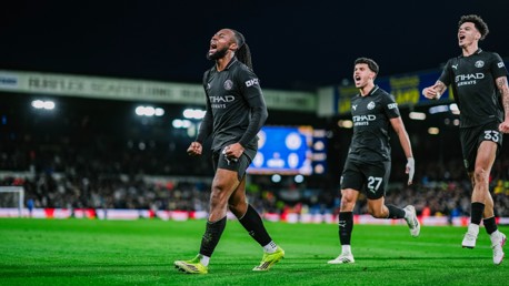 Three football players in black kits celebrating a goal on a soccer field. The stadium background includes visible sponsor logos and crowd.