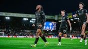 Three football players in black kits celebrating a goal on a soccer field. The stadium background includes visible sponsor logos and crowd.