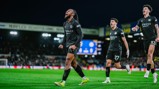Three football players in black kits celebrating a goal on a soccer field. The stadium background includes visible sponsor logos and crowd.