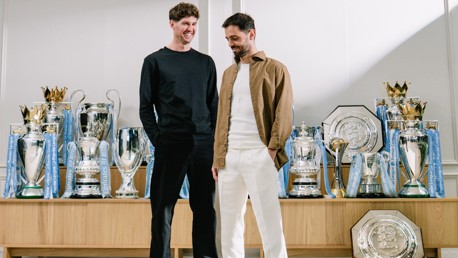 Two individuals stand in front of Manchester City's trophy collection, including Premier League, FA Cup, and other trophies.