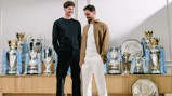 Two individuals stand in front of Manchester City's trophy collection, including Premier League, FA Cup, and other trophies.
