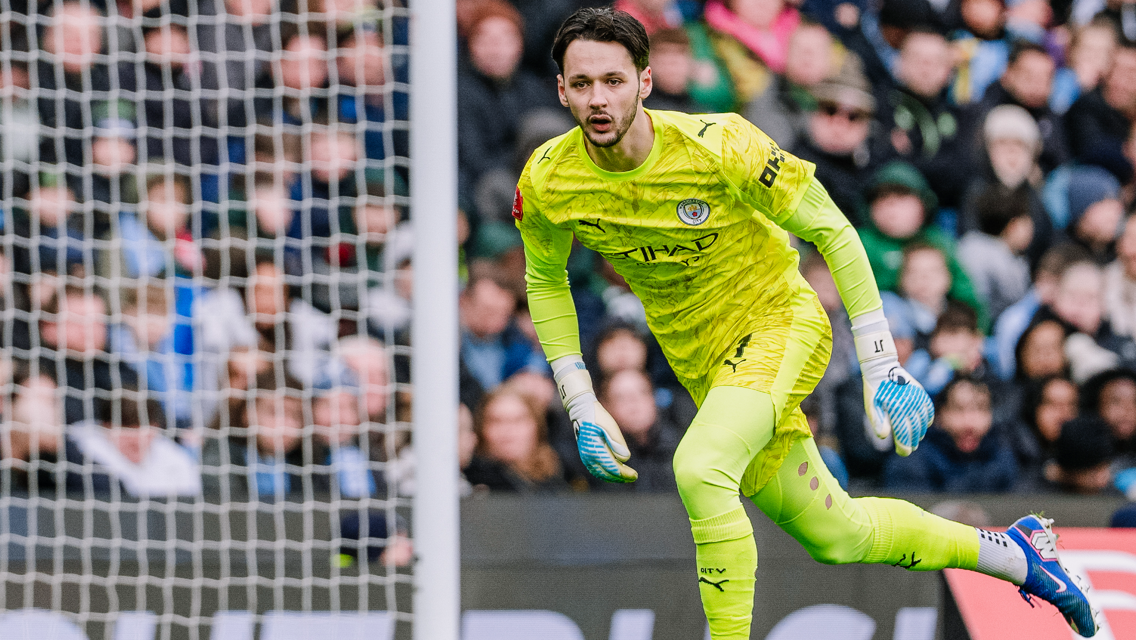 A soccer goalkeeper dressed in a bright yellow Manchester City uniform is seen in action, positioned near the goal net with a crowd in the background.