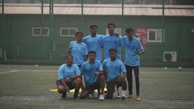 A group of youths wearing light blue sports jerseys with 'ETIHAD' logo, posing on a soccer field. The background includes a green fence and a building with a Coca-Cola vending machine.