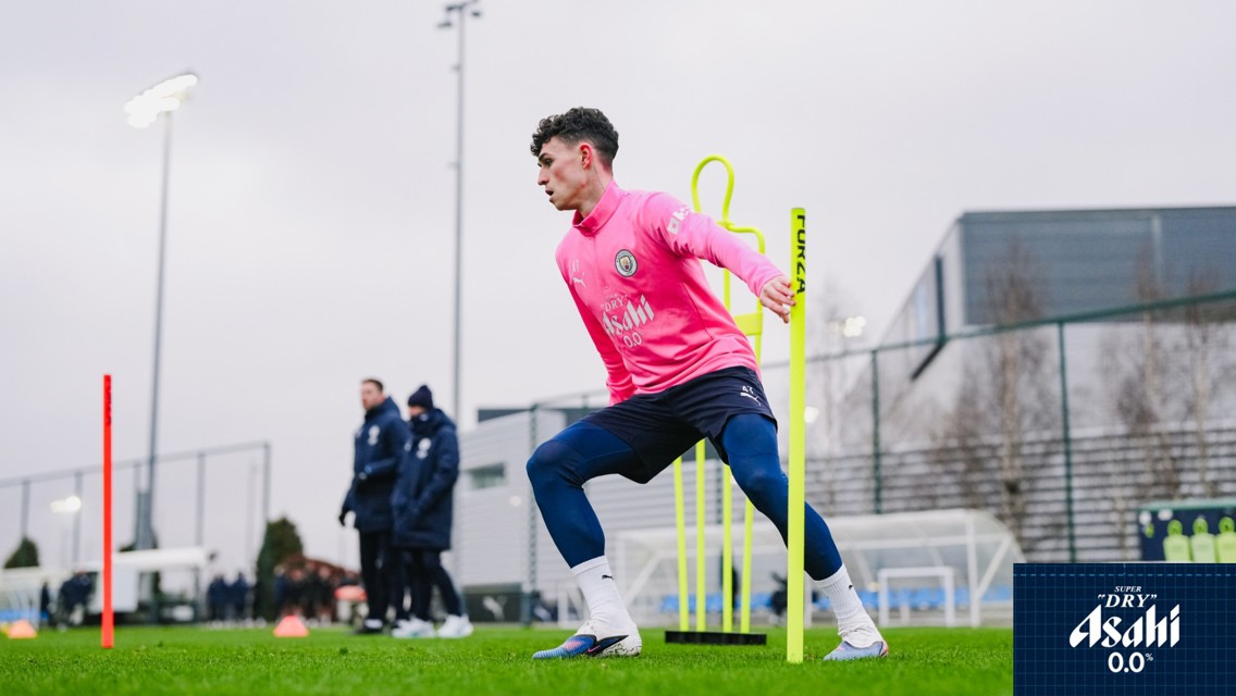 A football player in a pink Manchester City training kit practices agility exercises with a pole on a grassy field. Other team members are seen in the background, and the training ground includes sports facilities.