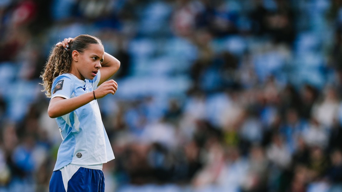 A woman soccer player on the field adjusting her ponytail, wearing a light blue and white jersey with the number 7. The background is blurred, showing a crowd in the stands.