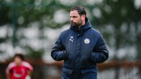 A Manchester City coach is wearing a Puma jacket with the club's logo while standing outdoors during a training session. The initials 'OR' are embroidered on the jacket.