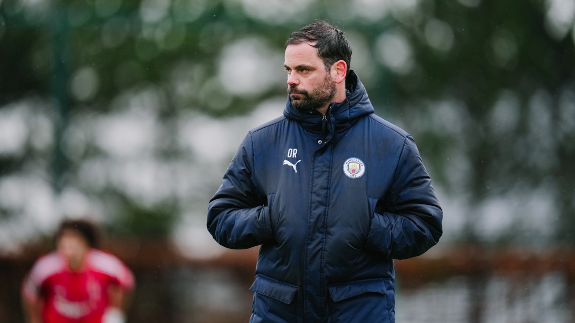 A Manchester City coach is wearing a Puma jacket with the club's logo while standing outdoors during a training session. The initials 'OR' are embroidered on the jacket.