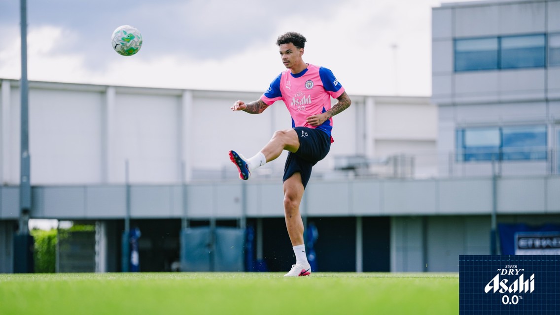 A football player in a pink training kit kicks a ball mid-air on a grassy field. The background shows a building and advertising signage.