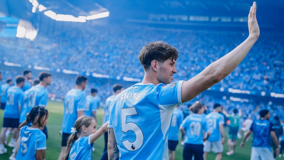 A football player with number 5 on jersey waves at a stadium packed with Manchester City fans. Other players and children in Manchester City kits can be seen in the background.