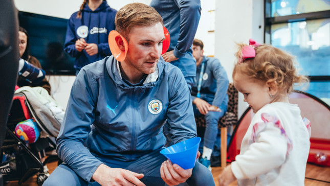 An individual wearing a Manchester City tracksuit kneels in front of a child holding a blue cone, with people standing and sitting around.
