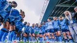 Manchester City women's team in a huddle, wearing light blue jerseys, preparing for a match. Shot taken from a low angle under a stadium roof.