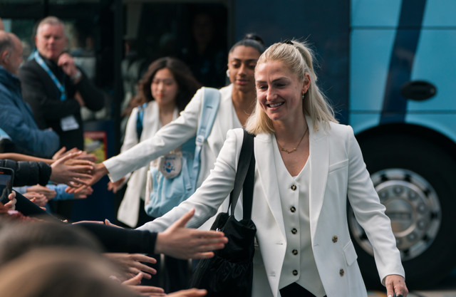A woman in a white jacket is walking past a group of people, extending her hand for a greeting. The background shows a blurred crowd and a bus.