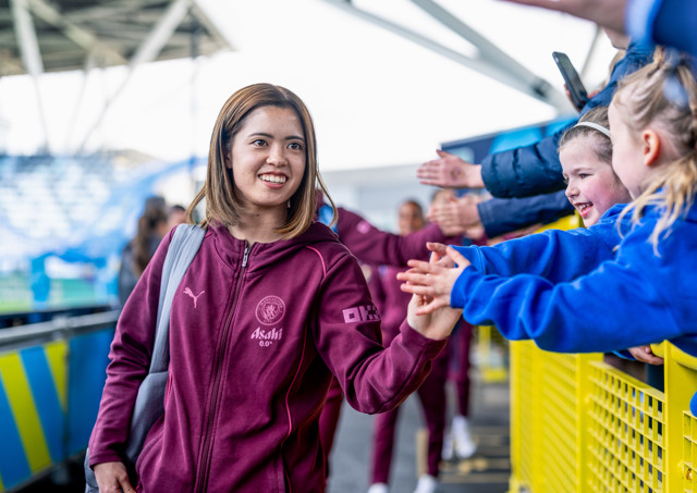 Manchester City female player in a maroon Puma tracksuit high-fives fans with one carrying a smartphone, in a stadium setting with blurred faces.
