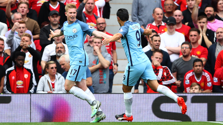 Two Manchester City players in light blue kits celebrate after scoring, with Manchester United fans in red kits reacting in the background.