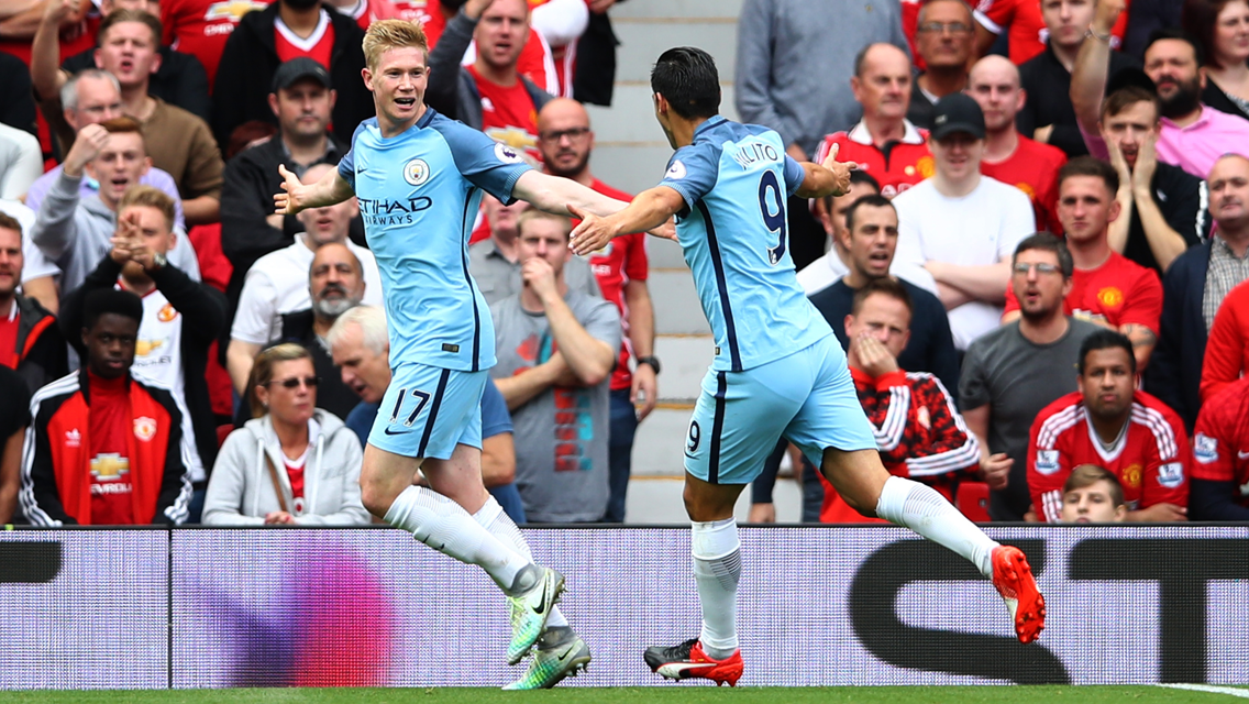 Two Manchester City players in light blue kits celebrate after scoring, with Manchester United fans in red kits reacting in the background.