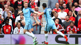 Two Manchester City players in light blue kits celebrate after scoring, with Manchester United fans in red kits reacting in the background.